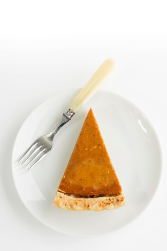 A piece of pumpkin pie, a sliced pastry ready to eat with a fork, on a white plate against a white background. The fresh, homemade baked dessert is a gourmet staple of an autumn Thanksgiving feast.
