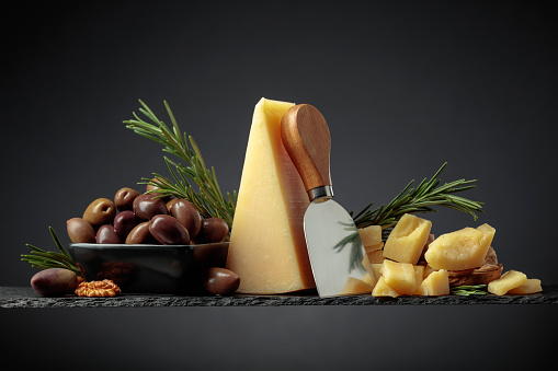 Parmesan cheese with knife, olives, and rosemary on a black background.