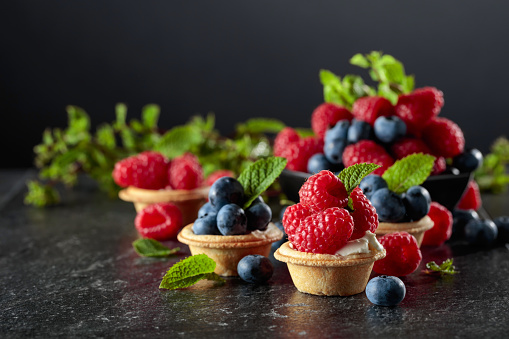 Small tartlets with fresh raspberries and blueberries garnished with mint on a black background.