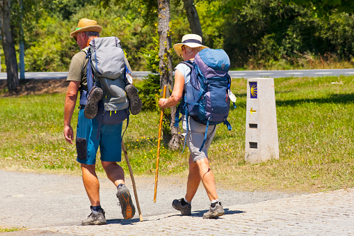 Senior couple walking, carrying their backpacks with scallops hanging, symbol of the pilgrimage, walking the camino de Santiago, ancient pilgrimage route .Directional milestone in the background.