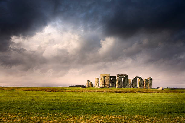 Distant overview of Stonehenge against stormy skies The sun pierces through a dense cloud layer to illuminate the iconic ancient temple of Stonehenge national trust photos stock pictures, royalty-free photos & images