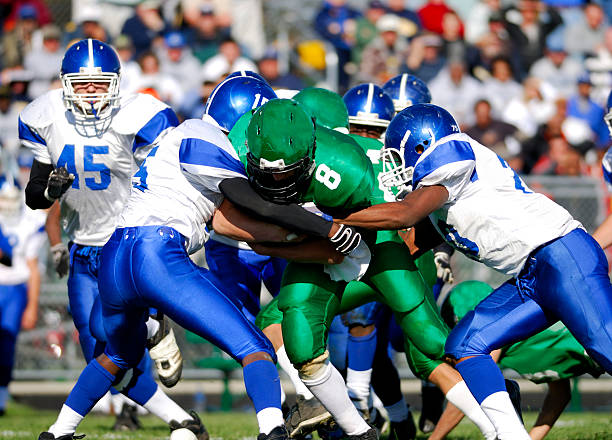 A football player is running with the ball in his arms.  He is holding the ball tight, while two players from the other team are trying to take it.  The man with the ball is dressed in a blue and white uniform.  The other team's uniform is black and yellow.