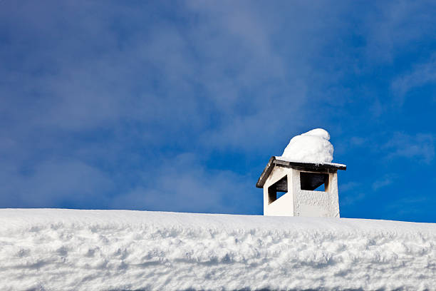 A chimney with a cap of snow stands on the snow-covered roof of a chalet against a sky background.