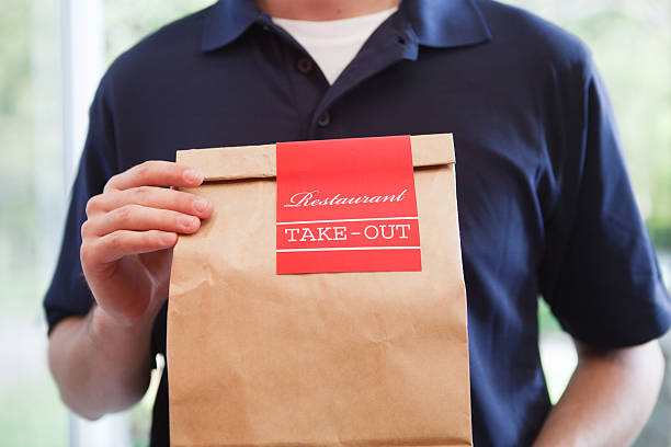 Take Out Food Delivery Person Holding Bag of Restaurant Dinner Close-up of an unidentifiable delivery man holding a take out food brown paper bag of restaurant dinner, delivering the fast food meal package. Waist up view of Caucasian delivery person wearing a blue polo shirt uniform and holding labeled container. The convenience service is used by USA homes or businesses. fast food uniform stock pictures, royalty-free photos & images