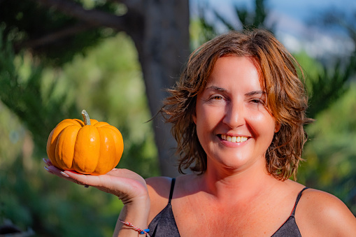 Smiling beautiful young woman looking at the camera and holding a natural fresh small pumpkin in her open hand. Sunny outdoor day with natural background and copy space. Happy Halloween concept.