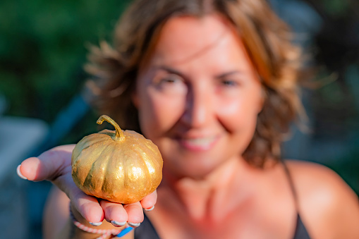 Smiling beautiful young woman holding a decorative golden small pumpkin in front of her face. Sunny outdoor day with nature background and copy space. Happy Halloween concept.