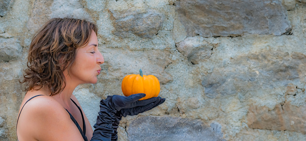 Beautiful young woman holding a natural fresh small pumpkin in her black gloved hands and preparing to kiss it. Sunny outdoor day with natural background and copy space. Happy Halloween concept.