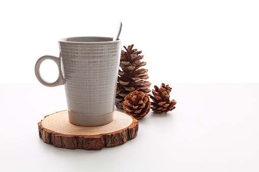 A gray ceramic mug with a spoon inside over a wooden coaster next to a a pine cones over a white table in front of a white background.