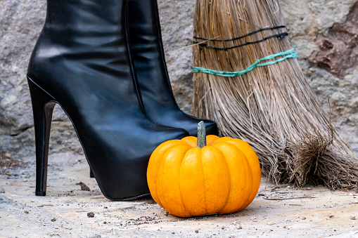 Beautiful Halloween decoration with black high heeled platform boots, witch broom and cute orange pumpkin on stone background for poster, greeting invitation, business banner. Outdoor photography with natural daylight and copy space. Side view.