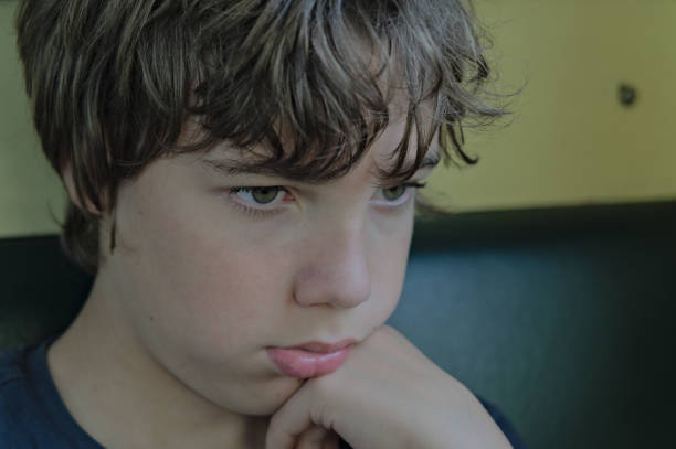A 12 year old boy in a blue polo shirt sits at a table, resting his head on his hands.