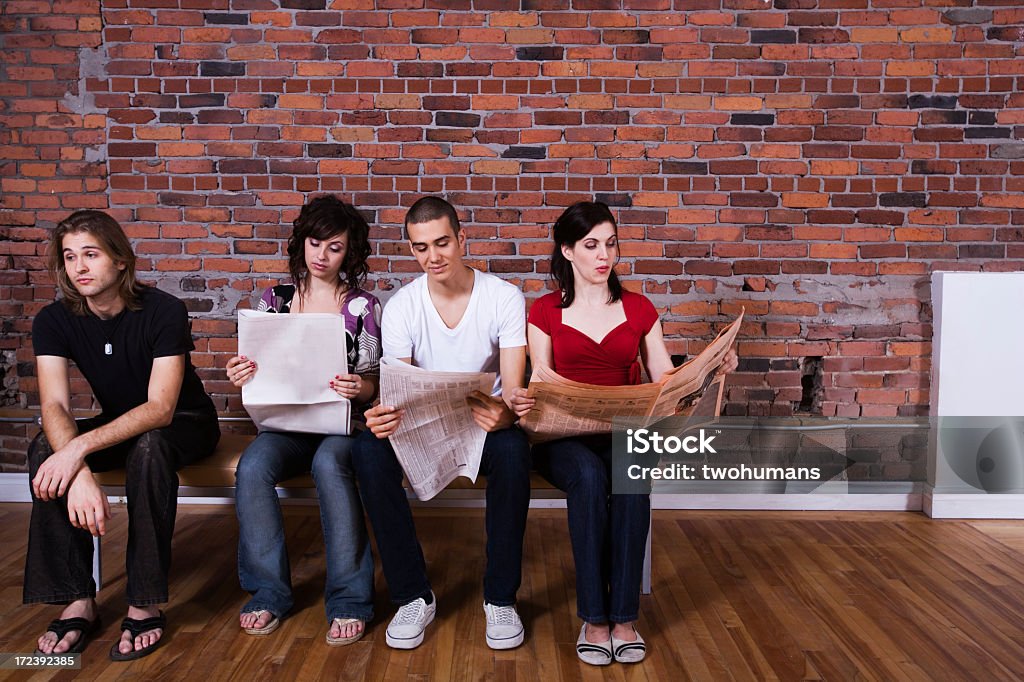 Friends sitting on a bench with three reading a newspaper Four persons sitting and reading the paper except one bored young man. Concepts : information, relationship; communication; exclusion.  Newspaper Stock Photo