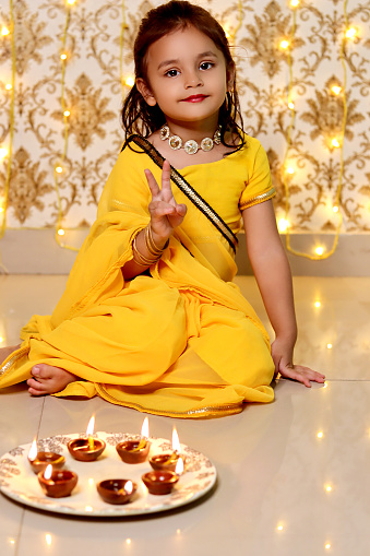 Small baby girl celebrating Diwali festival at home against light background. Diwali is the Hindu festival of lights with its variations also celebrated in other Indian religions. It symbolises the spiritual \