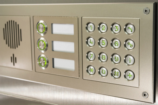 This is a angled close-up of a modern, front door security system or intercommunication system, with a dialing key pad, three calling buttons and a speaker for voice transmission. The whole setup is built from glossy aluminum, and the buttons are illuminated in green.