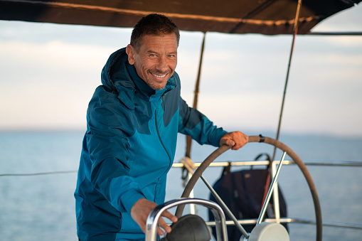 Man sailing on open water at sunset. He is standing over helm and wearing t-shirt with common sailor's pattern. Trolling fishing rode behind him