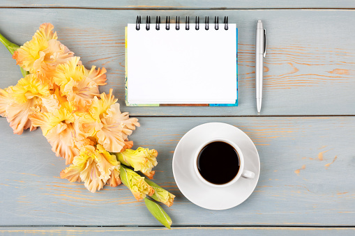 Artistic composition with opened blank notepad and pen, cup of black coffee, orange-yellow gladiolus flower on blue wooden table. Festive office desktop concept. Morning coffee cup.