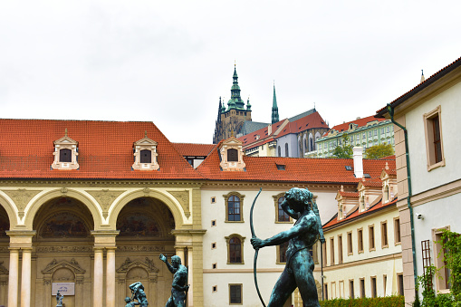 Ancient architectural building with arches, columns, beige walls and a red tiled roof and antique green statues in the foreground. Prague, Czech Republic, October 2022