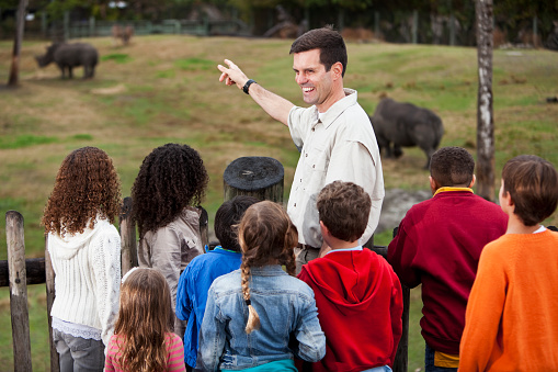 Zoo keeper (30s) with multi-ethnic group of elementary school children at zoo, standing on observation deck overlooking rhinoceros exhibit.