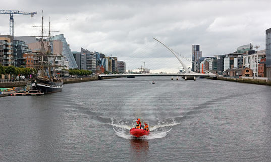 Dublin, Ireland - August 3, 2023 - Orange rubber boat travelling down the river Liffey on the Docklands on a cold overcast summer day, with the Samuel Beckett bridge in the backround