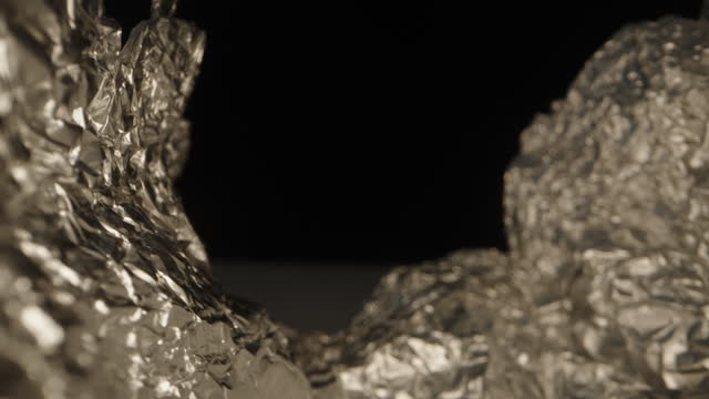 The camera moves along the creases of crumpled kitchen aluminum foil, highlighting its shiny texture against a black background. Macro shot.