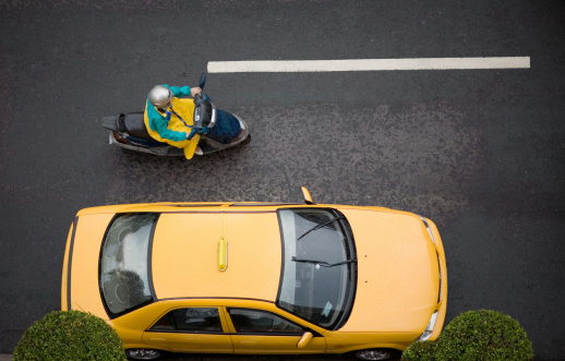 A close aerial view reveals a yellow cab being overtaken by a woman on a motorscooter. The black tar road provides a natural isolated background with plenty of copy space. Eos 5D. See more Asian images in