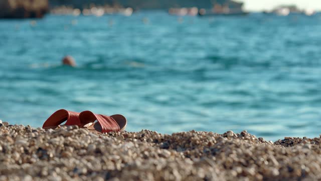 Red flip flop or sandal shoes on small pebbles beach, blurred calm turquoise sea with person swimming behind. Afternoon sunny holiday resort scene