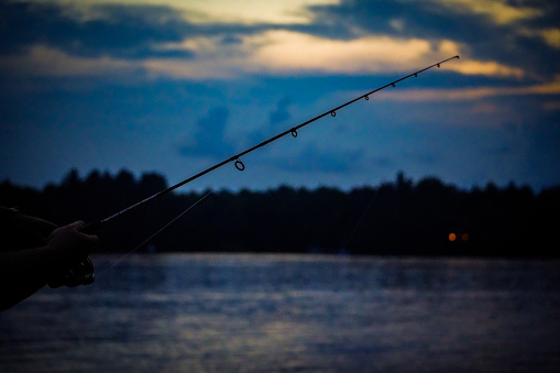 The sun sets through clouds in the distance as a fishing pole casts across a lake
