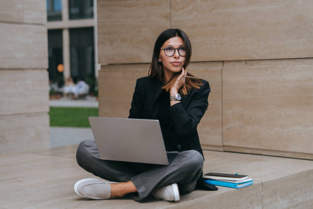 istock Pensive Hispanic young businesswoman in black jacket using laptop sitting outside looks up dreaming about new project. Mindful student girl in glasses prepares for exam. Business, education, success. 1724166364