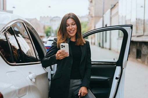 Brunette Hispanic businesswoman in black jacket standing at opened car holds phone smiles talks using video chat. Cheerful American female entrepreneur goes to meeting. Financial people.