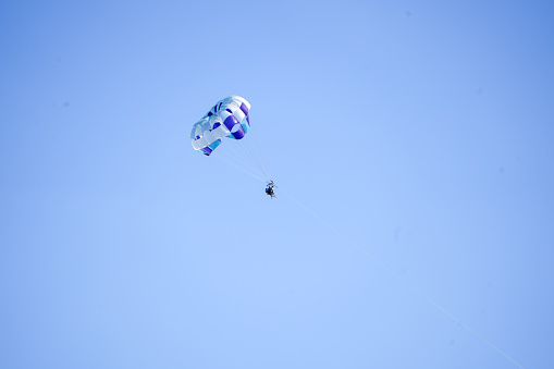 Purple, gray, and white parasail is pulled across a bright blue summer sky as tourists hang below