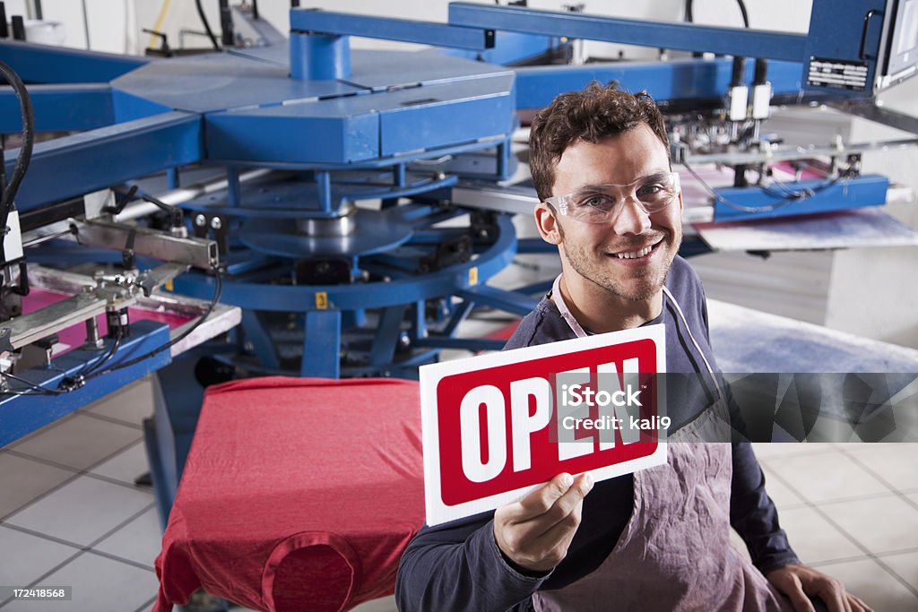 Man in screen printing business Small business - young man (20s) working in screen printing shop, holding OPEN sign. 20-29 Years Stock Photo