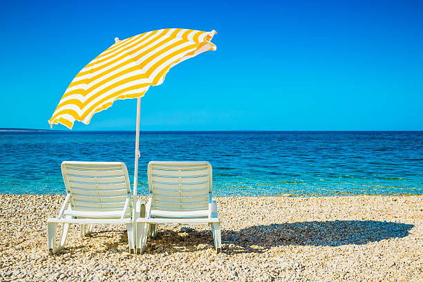 Sun loungers stand under an umbrella on a cobbled beach behind a fence overlooking the sea. High quality photo