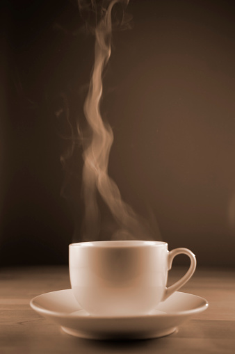 A sepia toned image of a plain white coffee cup in a saucer resting on a wooden table with steam rising from the cup. The cup in shot against a plain dark background with a shallow depth of field.  