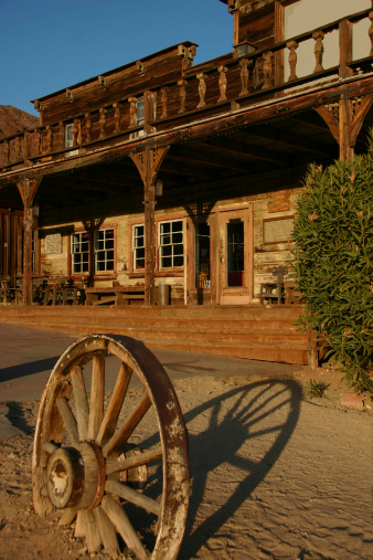 Front facade and porch of old west style building with wagonwheel in foreground and blue sky background.