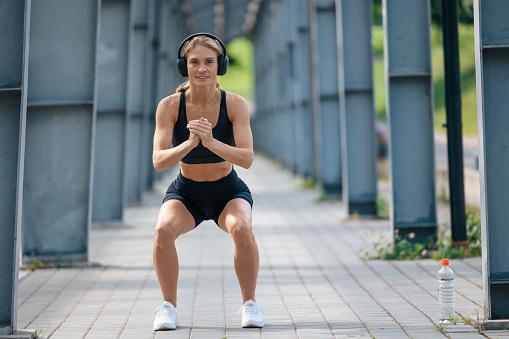 Sporty fit woman with headphones listening music squatting outdoor doing fitness exercises in summer morning.