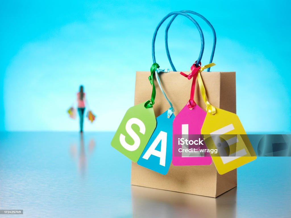 Carrying Shopping Bags with the Word Sale in Multi Colours "Multi coloured labels spelling the word sale hanging from a brown paper carrier bag on a stainless steel background, with blue lighting, copy space and a female in the background carrying shopping bags.Click on the link below to see more of my retail images." Advertisement Stock Photo