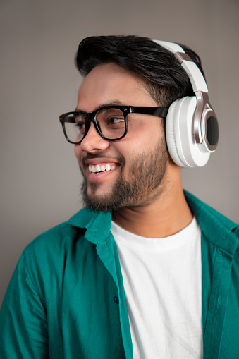 Bespectacled relaxed, happy indian young man standing against the isolated gray background and listening music through headphones.