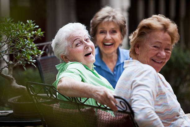 Three senior women (80s) sitting on patio, talking and laughing together.  Focus on woman in green shirt.