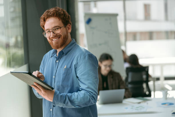 istock Positive bearded male manager is using digital tablet standing in modern coworking and looks camera 1724406123