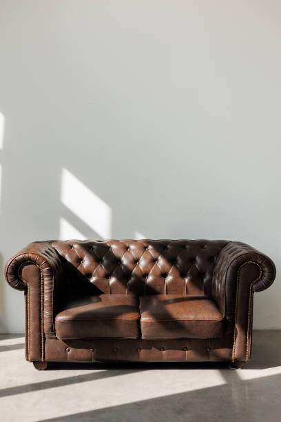 istock Vintage quilted classic brown sofa against white wall with sunlights from window on concrete floor at studio. Natural leather with rhombic stitching with buttons. Furniture design. Copy space 1724408816