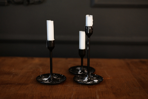 Modern three black glass candlesticks with white candles on wooden table in antique style on dark wall background. Minimalist elegant decor with candle holders with wax different size