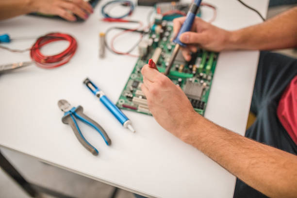 Male student sitting at desk in classroom using soldering iron and tweezers while learning to repair computer parts
