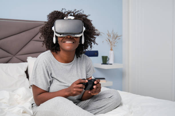 African young woman playing video games with a VR headset sitting on bed. African young woman playing video games with a VR headset sitting on bed in the morning. world cyber games photos stock pictures, royalty-free photos & images