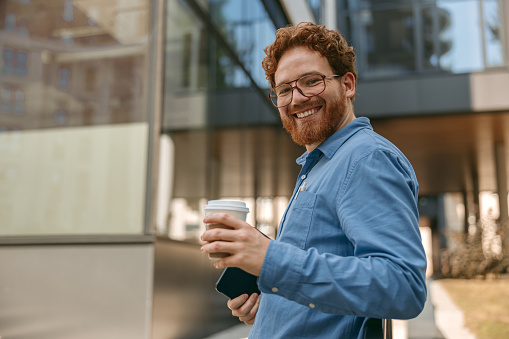 Bearded smiling businessman with digital tablet is drinking coffee during break outside of office