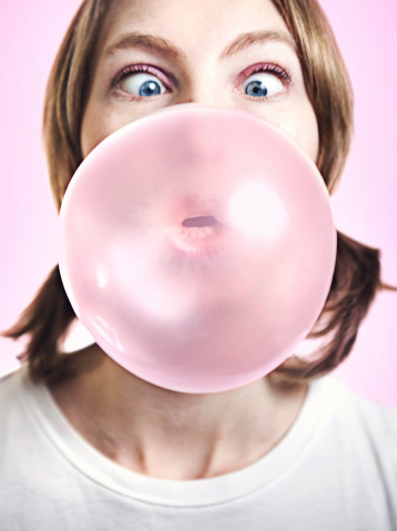 A large pink bubble gum bubble obscures a young woman's face, her eyes crossing to take in how big it is.  Vertical portrait on pink background.  Focus on bubble and lips.