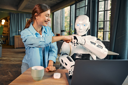 Young woman with a cup of coffee and a humanoid robot working while sitting at a laptop in a modern office, fist to fist gesture. Collaboration between humans and artificial intelligence.