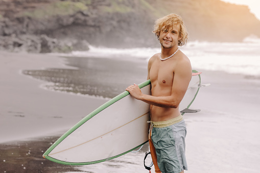 Handsome fit young blond man with mock up surfboard waits for wave to surf spot at sea ocean beach with black sand and looks at camera. Concept of sport, fitness, freedom, happiness, new modern life.