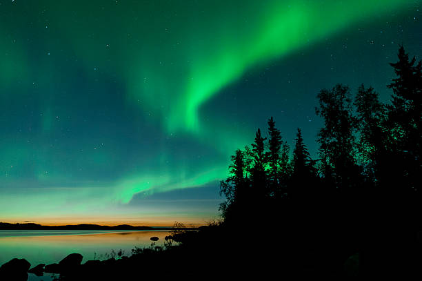 Summer aurora on lake Summer aurora at Cassidy Point, Northwest Territories, Canada with sun setting in background and silhouette of trees in foreground. aurora borealis stock pictures, royalty-free photos & images