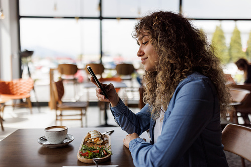 Young smiling woman using mobile phone while having breakfast at a cafe