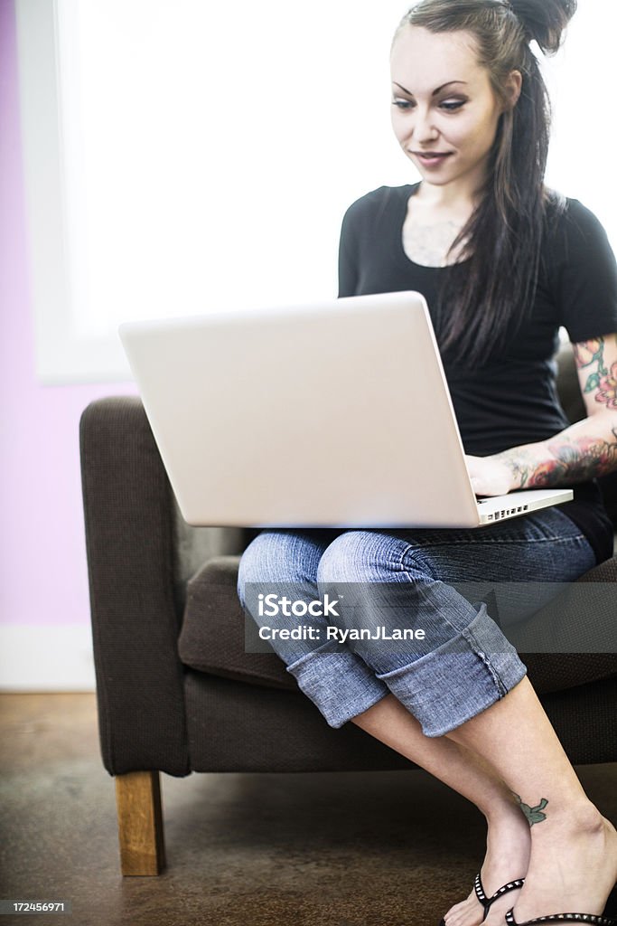 Tattoed Young Woman on Laptop A beautiful young adult woman with arm tattoos and sleeve sits in a comfy chair in her bright studio apartment, working on her laptop computer.  Bright sunlight shines in through the large bay windows behind her. 20-29 Years Stock Photo