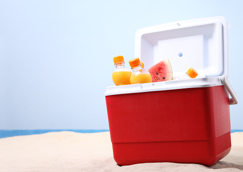 Open ice box with orange juice bottles on white sand at the beach.